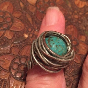 May include: A handcrafted ring featuring a vibrant turquoise stone, encased in a woven wire design. The ring's metal has a rustic, aged appearance, adding to its unique, bohemian style. The turquoise stone is a striking focal point.