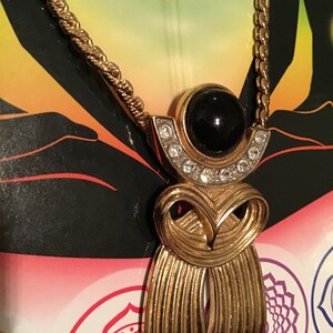 Puede incluir: Un collar de cadena dorada con un colgante que presenta una piedra negra rodeada de diamantes de imitaci&oacute;n transparentes. El colgante tiene la forma de una figura estilizada con un dise&ntilde;o fluido.