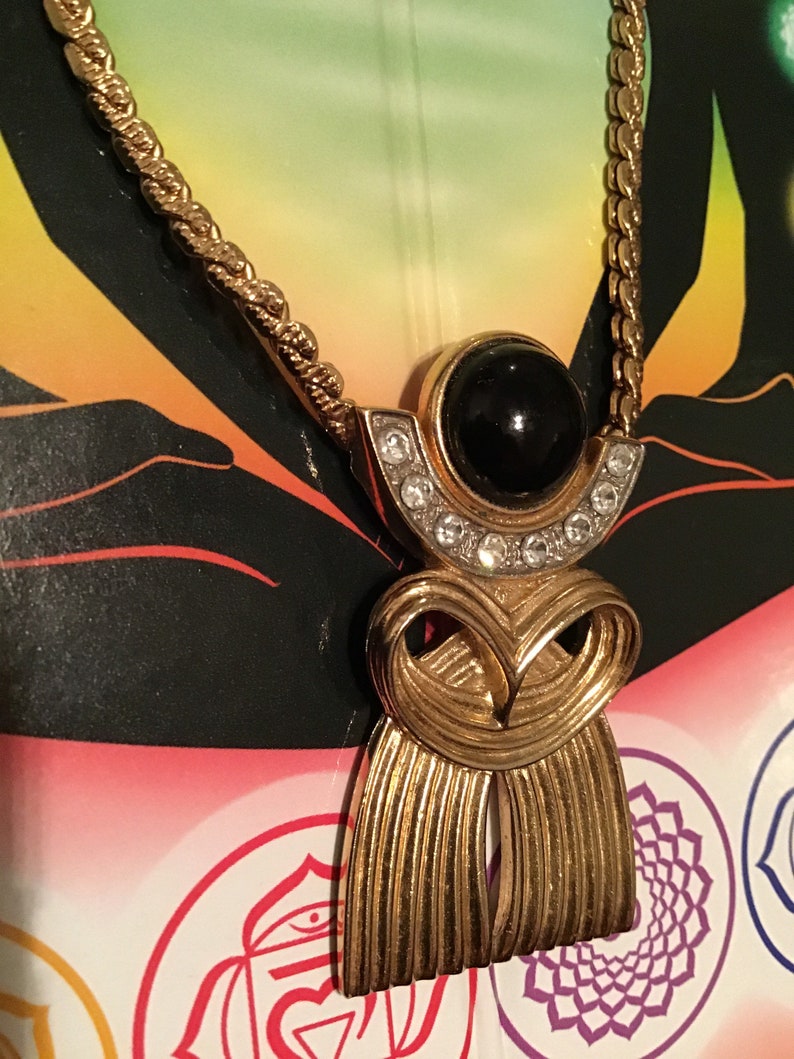 Puede incluir: Un collar de cadena dorada con un colgante que presenta una piedra negra rodeada de diamantes de imitaci&oacute;n transparentes. El colgante tiene la forma de una figura estilizada con un dise&ntilde;o fluido.