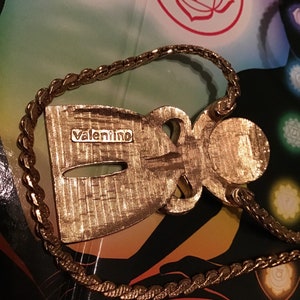 Puede incluir: Un collar con un colgante en forma de figura estilizada de color dorado, con la palabra "valentino" inscrita en la parte posterior. El collar tiene una cadena gruesa y trenzada.