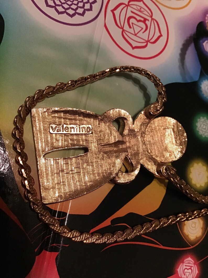 Puede incluir: Un collar con un colgante en forma de figura estilizada de color dorado, con la palabra "valentino" inscrita en la parte posterior. El collar tiene una cadena gruesa y trenzada.