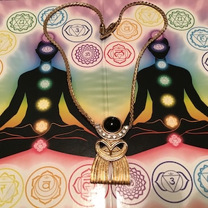 Puede incluir: Un collar de cadena dorada con un gran colgante que presenta una piedra negra y diamantes de imitaci&oacute;n. El colgante tiene forma de figura estilizada con un fondo tipo flecos. El collar se muestra sobre un fondo con una imagen colorida de una persona en una postura meditativa con siete chakras.