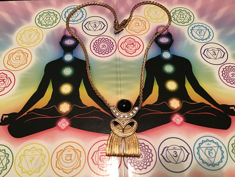 Puede incluir: Un collar de cadena dorada con un gran colgante que presenta una piedra negra y diamantes de imitaci&oacute;n. El colgante tiene forma de figura estilizada con un fondo tipo flecos. El collar se muestra sobre un fondo con una imagen colorida de una persona en una postura meditativa con siete chakras.