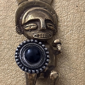 May include: A bronze-toned figurine of a stylized figure with a headdress and a shield with a black center. The figure is holding a small object in its right hand. The figurine is set against a textured, gold-colored background.