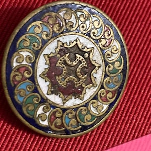 May include: A round, ornate pin with a gold-colored metal border. The pin features a central design with a star-like shape in red and gold, surrounded by a white circle and a blue border with colorful swirls. The pin is set against a red background.