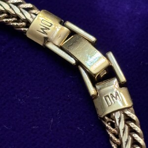 May include: Close-up of a gold-toned chain necklace with a woven design. The clasp is visible, featuring the initials "DM" engraved on it. The necklace is set against a dark blue background.