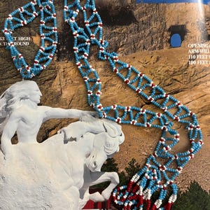 May include: A beaded necklace with a geometric design. The necklace features light blue beads with white and red accents. The necklace is displayed on a magazine page with text and a sculpture of a horse.