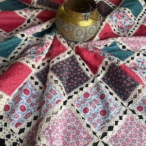 May include: A patchwork quilt with a floral pattern in shades of pink, teal, and dark green. The quilt has a crocheted border and a decorative brass pot in the center. The quilt is a mix of floral and geometric patterns.