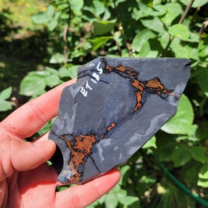 May include: A flat, irregularly shaped piece of dark gray stone featuring prominent, branching veins of rusty orange and brown mineral deposits. The surface of the stone is marked with the white handwritten text ET185 in the upper left corner.