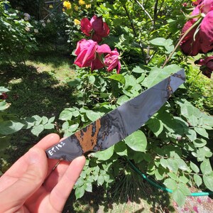 May include: A flat, tapered knife blade blank featuring a marbled pattern of dark gray, black, and metallic copper tones. The handle area is marked with the white handwritten text ET182, showcasing a distinct contrast between the dark stone-like finish.