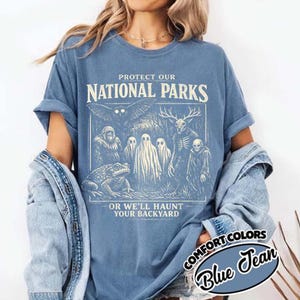 Peut inclure: T-shirt bleu avec un motif qui dit "PROTECT OUR NATIONAL PARKS" et "OR WE'LL HAUNT YOUR BACKYARD." Le motif présente diverses créatures effrayantes. Le t-shirt est étiqueté "COMFORT COLORS Blue Jean."