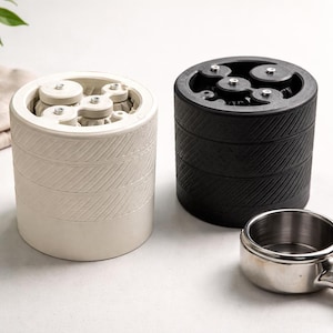 May include: Two cylindrical coffee grinders, one cream-colored and one black, with a textured diagonal pattern. The cream grinder has visible internal gears. A stainless steel portafilter with a wooden handle is in the foreground. Coffee beans are scattered nearby.