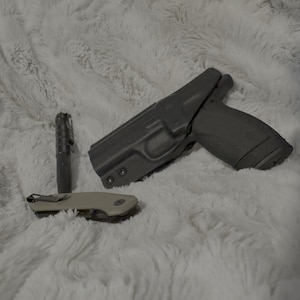 May include: A collection of tactical gear including a handgun in a holster, a small flashlight, and a folding knife. The items are arranged on a soft, white, textured surface.
