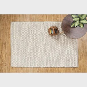 May include: A cream-coloured, textured area rug with a woven design. The rug is rectangular and sits on a wooden floor. A small, round, dark brown table with a plant and a decorative basket are visible.