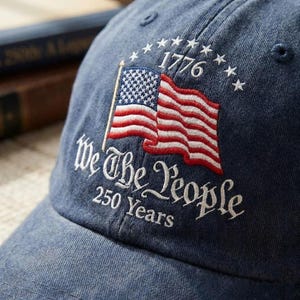 以下が含まれることがあります： 青い野球帽。刺繍されたアメリカ国旗、テキスト「1776」と「We The People 250 Years」が白でデザインされています。デニムのような素材で作られています。