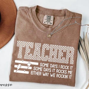 以下が含まれることがあります： エスプレッソ色のTシャツ。前面に市松模様の「TEACHER」の文字と「SOME DAYS I ROCK IT, SOME DAYS IT ROCKS ME, EITHER WAY WE ROCKIN' IT」のテキストがプリントされています。シルバーのネックレスがTシャツの上に置かれています。