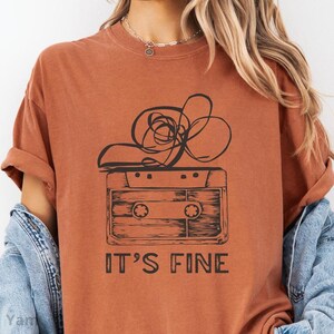 以下が含まれることがあります： 黒いカセットテープと抽象的な線が描かれた、焦げたオレンジ色のTシャツ。「IT'S FINE」の文字が太字でプリントされています。Tシャツは半袖でゆったりとしたフィット感です。モデルはデニムジャケットを着ています。