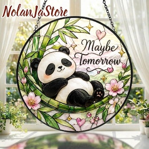 Op de afbeelding: Een ronde glas-in-lood zonnevanger met een slapende panda op groene bamboestengels. Het ontwerp bevat roze kersenbloesems, kleine hartjes en de tekst "Maybe Tomorrow" in zwart handschrift, gevat in een zwart metalen frame.