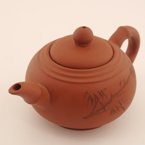 Op de afbeelding: Roodbruine keramische theepot met een afgeronde body, een deksel met een kleine knop en een gebogen handvat. De theepot heeft een tuit en is versierd met een bamboetakontwerp. Ontworpen voor het zetten van thee.