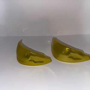 May include: Two curved, semi-transparent yellow plastic lenses with a smooth, glossy finish. These crescent-shaped components feature a distinct molded contour and are positioned side-by-side against a plain, neutral white background.