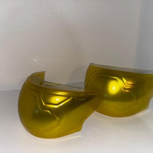 May include: Two curved, metallic gold shoulder armor pieces featuring a molded, geometric panel design. Each piece has a smooth, reflective surface with raised ridges and a contoured shape designed to fit over a shoulder area.