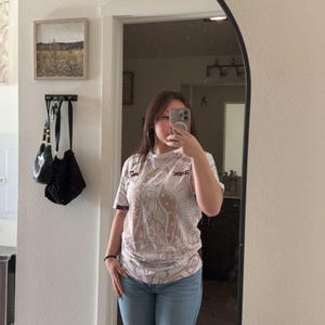 May include: A person wearing a white and brown graphic t-shirt with a design and the text "REK", paired with light blue ripped jeans. The person is standing in front of a mirror.