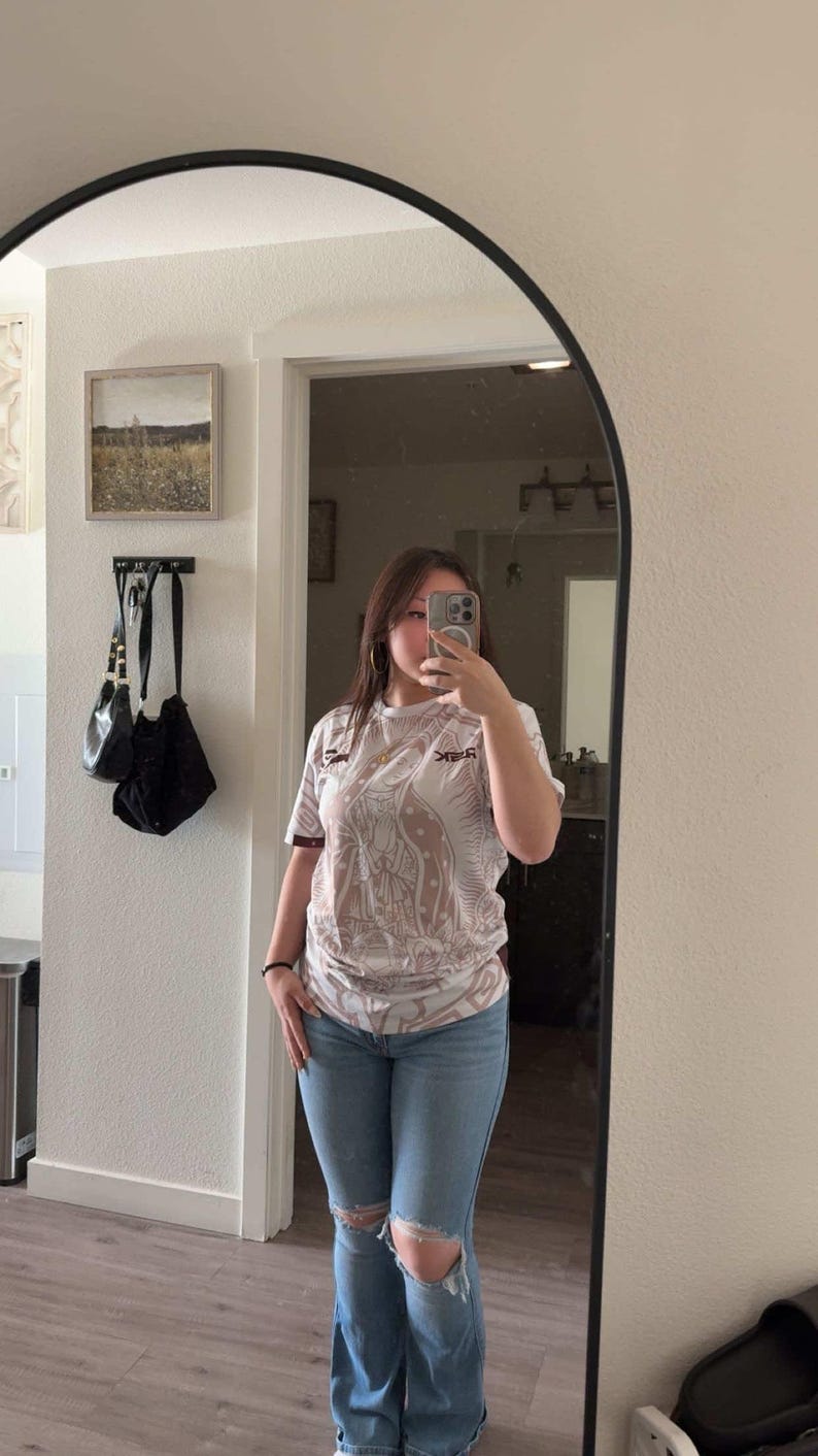 May include: A person wearing a white and brown graphic t-shirt with a design and the text "REK", paired with light blue ripped jeans. The person is standing in front of a mirror.