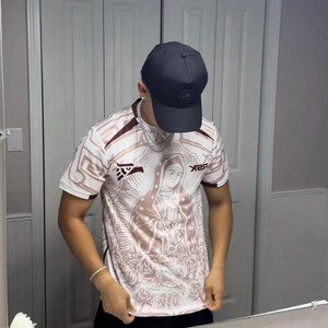 May include: A white and maroon short-sleeve jersey with a religious image on the front. The jersey has a crew neck and the text "X2R" on the right side. A black baseball cap is worn.