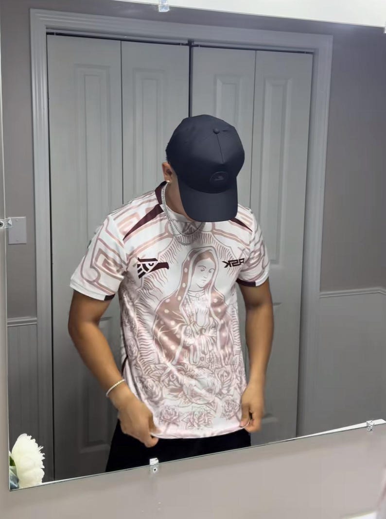 May include: A white and maroon short-sleeve jersey with a religious image on the front. The jersey has a crew neck and the text "X2R" on the right side. A black baseball cap is worn.