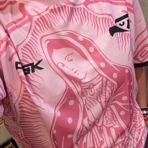 May include: Pink t-shirt featuring a detailed graphic of a religious figure with a halo, hands clasped in prayer, and floral accents. The shirt has a crew neck and short sleeves. The letters "FGK" and a stylized eagle are visible on the shirt.