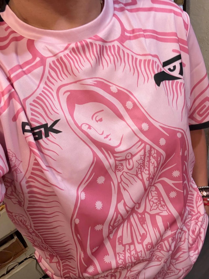 May include: Pink t-shirt featuring a detailed graphic of a religious figure with a halo, hands clasped in prayer, and floral accents. The shirt has a crew neck and short sleeves. The letters "FGK" and a stylized eagle are visible on the shirt.