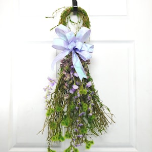 May include: A decorative door wreath featuring a moss-covered ring, a large iridescent lavender bow, and a cascade of dried flowers and greenery. The arrangement includes purple and green accents, creating a natural and elegant aesthetic.