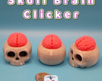 May include: Three beige skull-shaped clickers with bright pink brain-shaped tops. The text 'Skull Brain Clicker' appears above. A US quarter coin is placed near the objects for scale, alongside a small square logo reading 'Creatively Cubic'.