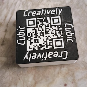 May include: A black and white QR code on a rectangular block. The word "Creatively" is printed in white at the top and bottom, with "Cubic" on the sides. The block is resting on a light-colored surface.