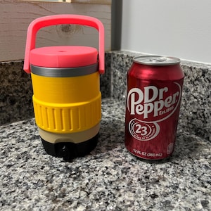 May include: A yellow and black pencil-shaped tumbler with a pink handle and lid, next to a red Dr. Pepper can. The can has white text and the number 23 in a circle. The tumbler is on a speckled gray surface.