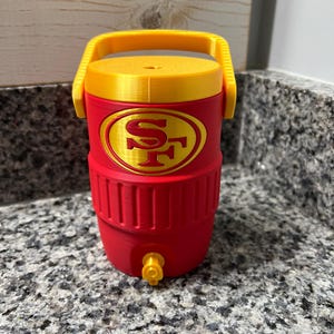 May include: A red and gold water cooler with a yellow handle and lid. The cooler features the San Francisco 49ers logo in gold. It has a tap at the bottom for dispensing liquids. The cooler is on a granite countertop.