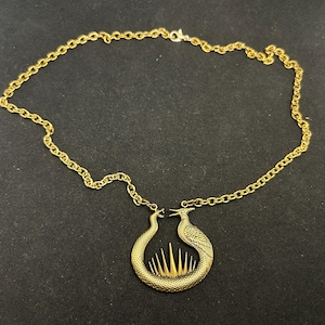 Puede incluir: Un collar dorado con un colgante que representa dos figuras de pájaros enfrentadas, con una base en forma de serpiente y un diseño de rayos de sol. El collar tiene una cadena delicada.