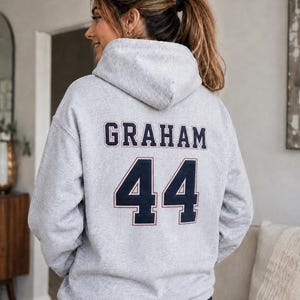 Op de afbeelding: Een lichtgrijze hoodie met de naam GRAHAM in donkerblauwe blokletters met een dunne rode omlijning, geplaatst boven het nummer 44 in hetzelfde donkerblauwe en rode kleurenschema, bedrukt op de achterkant van het kledingstuk.
