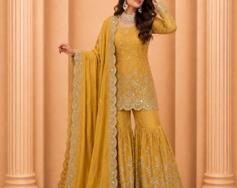 May include: Mustard yellow embroidered Sharara suit. The outfit includes a long-sleeved tunic, flared pants, and a matching scarf. The ensemble features intricate gold embroidery and a scalloped trim. The background is a peach color with architectural columns.