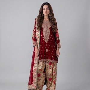 Op de afbeelding: Een bordeauxrode salwar kameez-ensemble met ingewikkelde gouden borduursels op het lijfje en de mouwen. De outfit bevat een bijpassende broek met een bloemmotief in beige en rood, en een doorschijnende bordeauxrode sjaal.