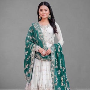 May include: A white and green embroidered Anarkali suit. The long-sleeved dress features intricate floral embroidery on the bodice and hem. A matching green dupatta with floral patterns drapes over the shoulders. The outfit is completed with embellished sandals.