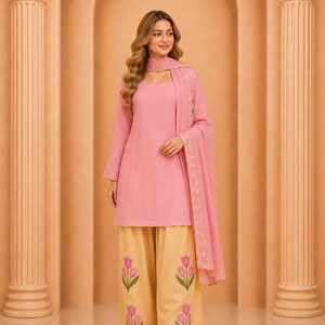 Puede incluir: Una túnica rosa de manga larga con escote cuadrado, combinada con pantalones beige de pierna ancha con motivos de tulipanes rosas bordados. Un dupatta rosa a juego con delicados bordados festoneados completa el conjunto.