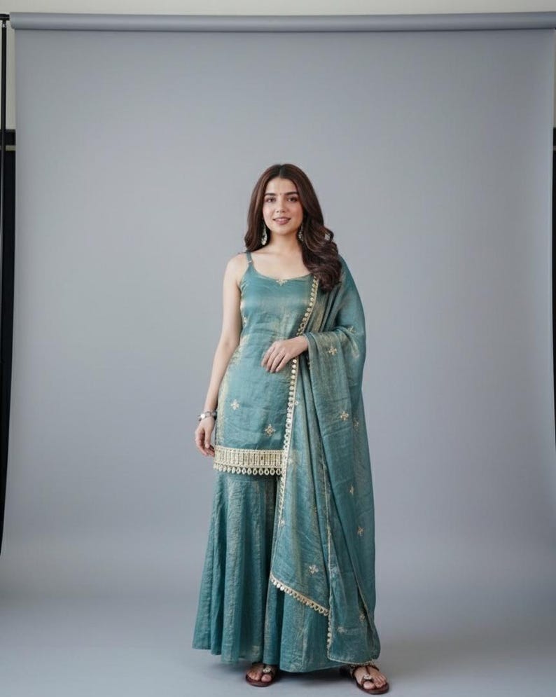 May include: A teal sleeveless kurta with thin straps and a matching flared sharara pant set. The outfit features gold embroidery, a decorative gold border at the hem, and a coordinating sheer teal dupatta with gold accents draped over one shoulder.