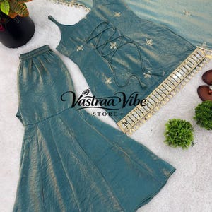 May include: A teal sleeveless top with a lace-up front, matching flared pants, and a coordinating dupatta. The set features gold embroidery, mirror work trim, and the text Vastraa Vibe Store. The fabric has a subtle metallic sheen throughout.