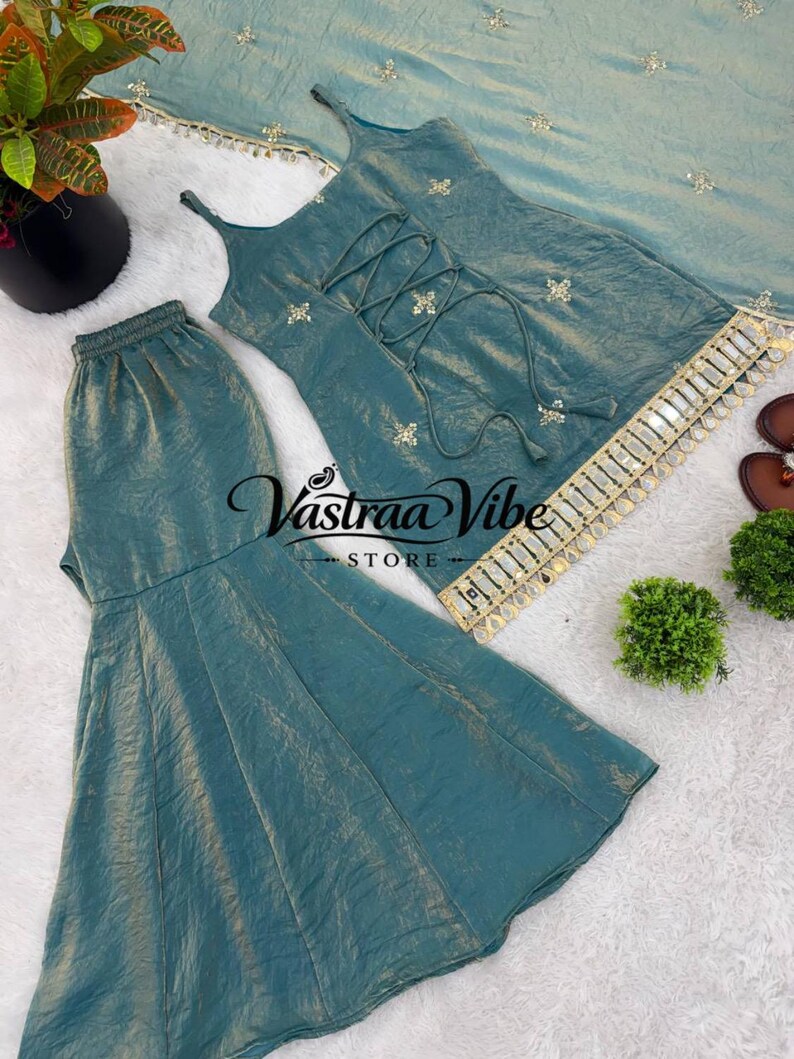 May include: A teal sleeveless top with a lace-up front, matching flared pants, and a coordinating dupatta. The set features gold embroidery, mirror work trim, and the text Vastraa Vibe Store. The fabric has a subtle metallic sheen throughout.