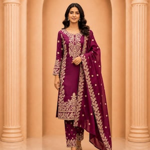 May include: A deep magenta long-sleeved tunic and matching trousers set featuring intricate gold embroidery along the neckline, cuffs, and hem. A coordinating sheer magenta dupatta with gold floral embroidery is draped over one shoulder.