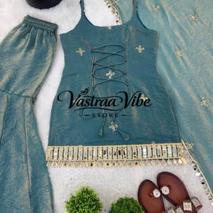 May include: A teal sleeveless top with a lace-up front, gold embroidery, and a mirrored fringe hem, paired with matching wide-leg pants and a dupatta. The top features the text Vastraa Vibe STORE in black script across the center chest area.