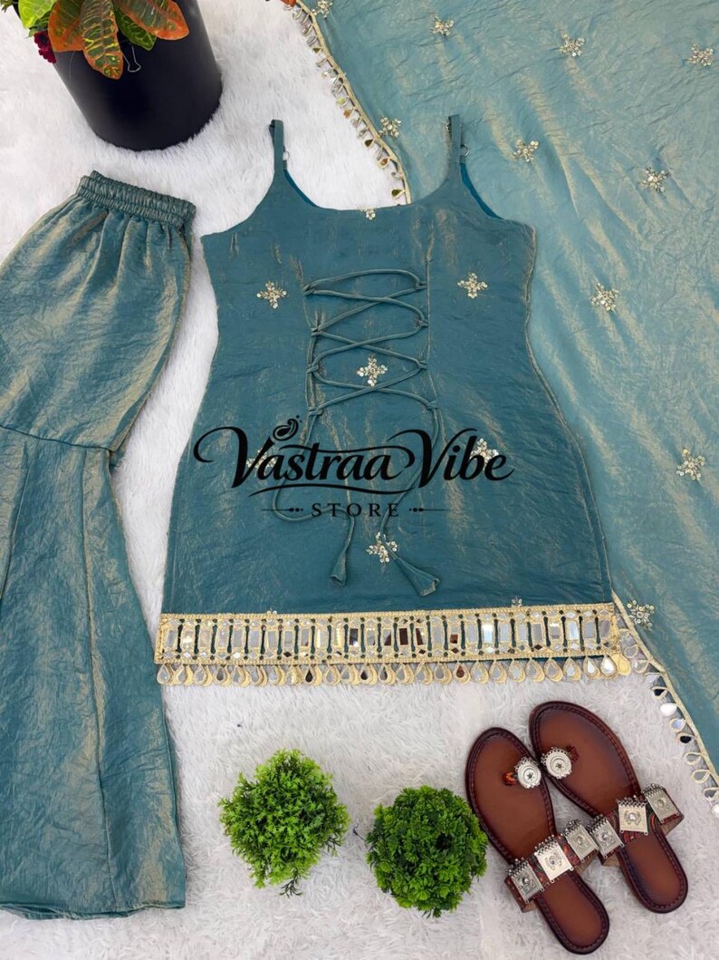 May include: A teal sleeveless top with a lace-up front, gold embroidery, and a mirrored fringe hem, paired with matching wide-leg pants and a dupatta. The top features the text Vastraa Vibe STORE in black script across the center chest area.