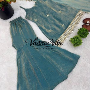 May include: A teal sleeveless top with thin straps and a matching flared skirt, both featuring a subtle metallic sheen. The top is adorned with small floral embroidery and a decorative fringe trim along the hem. The text Vastraa Vibe STORE is visible.