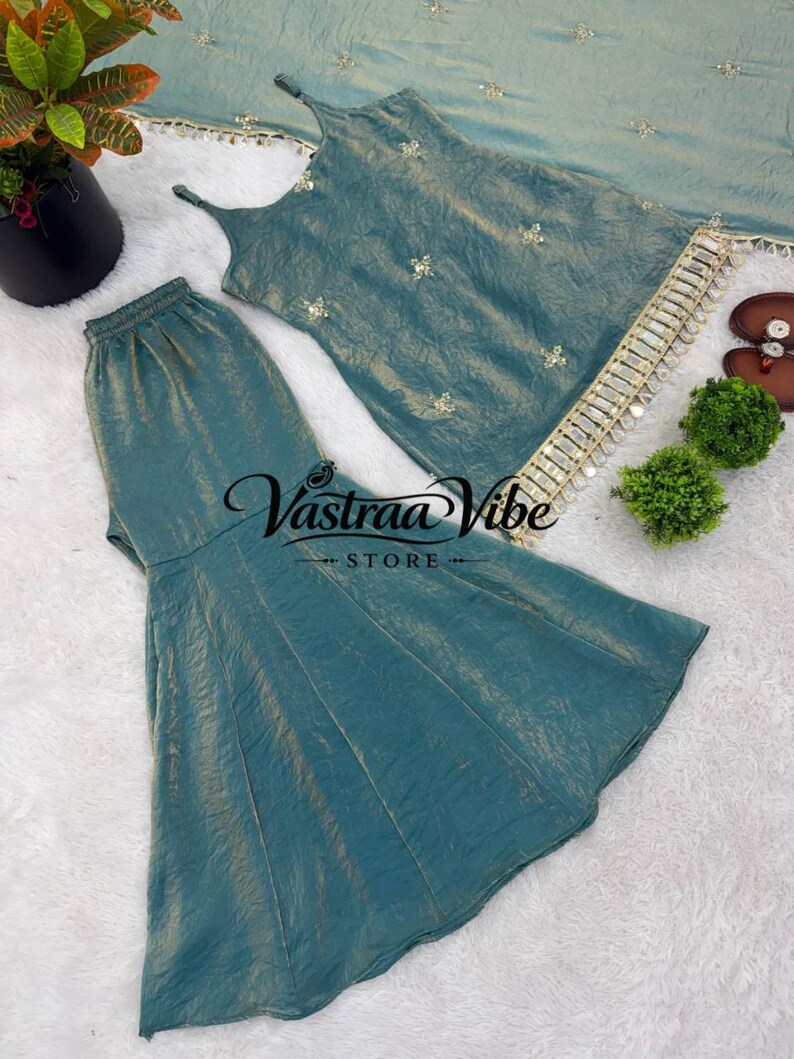 May include: A teal sleeveless top with thin straps and a matching flared skirt, both featuring a subtle metallic sheen. The top is adorned with small floral embroidery and a decorative fringe trim along the hem. The text Vastraa Vibe STORE is visible.